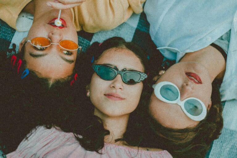 Three stylish women lying outdoors, wearing sunglasses and enjoying a relaxed moment together.